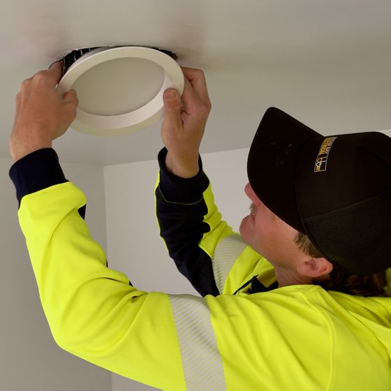 Two hands pressing a round white downlight into a ceiling, with the fitter in a hi-vis hoodie and black cap looking up at it